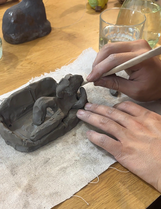 Close-up of hands sculpting a small clay figure on a beige cloth, with a finished clay sculpture in the background