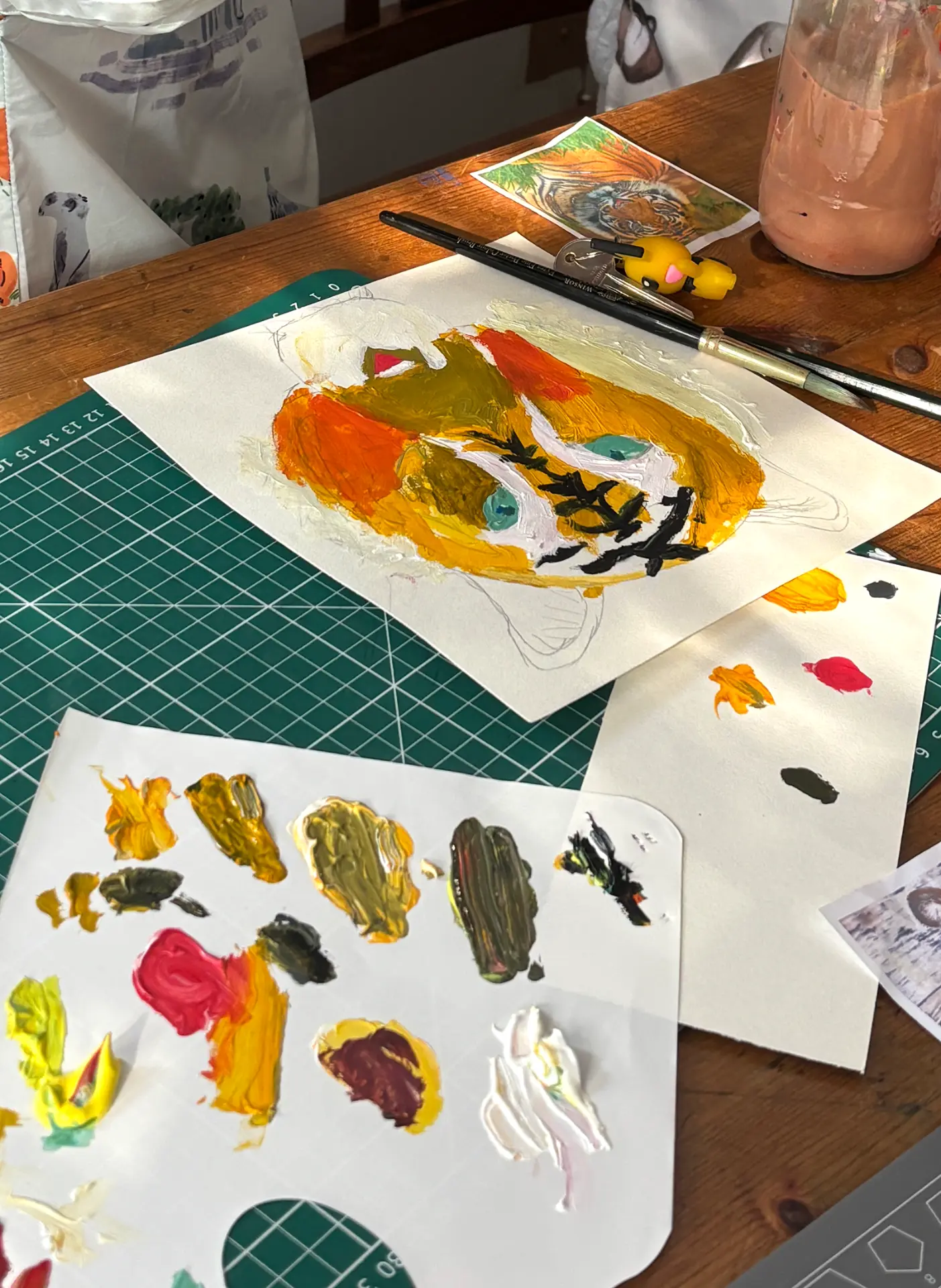 Top-down view of a wooden studio workspace with a tiger painting in progress, a paint palette, brushes and a jar of water